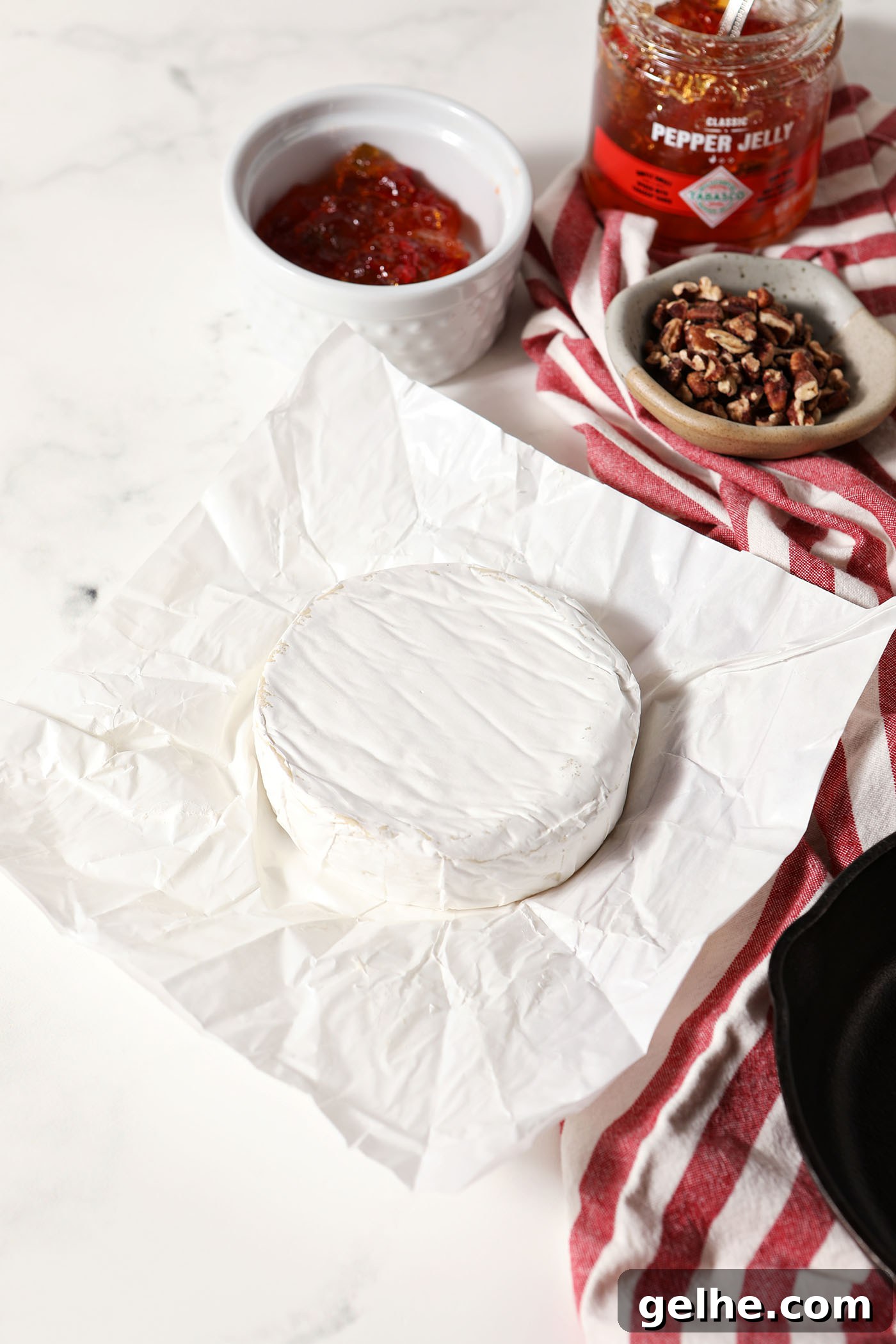 A wheel of brie next to bowls of pepper jelly and toasted pecans on a marble surface next to a red and white striped linen