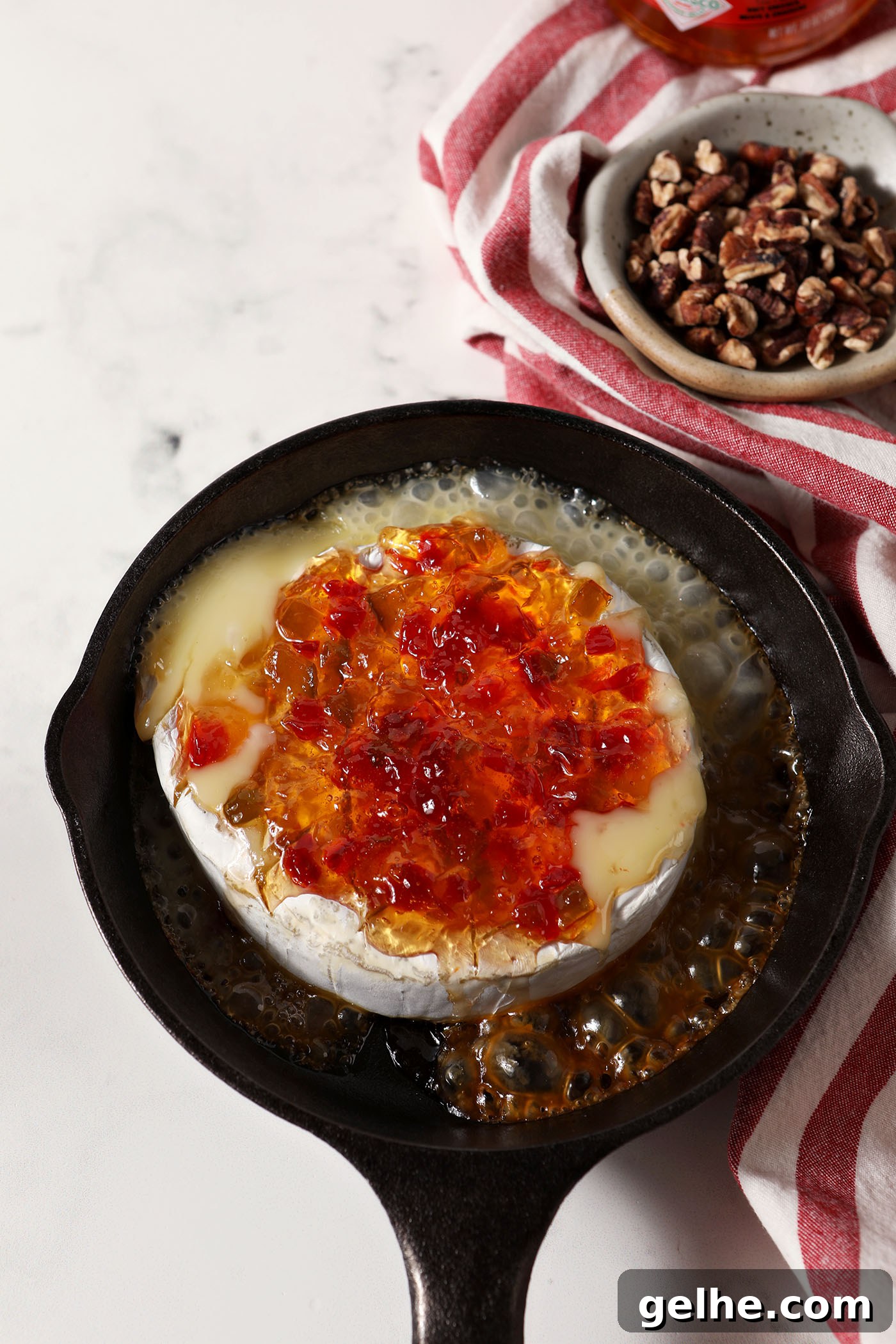 Overhead of a cast iron skillet holding a melty wheel of brie topped with pepper jelly on a marble surface next to a red and white striped linen