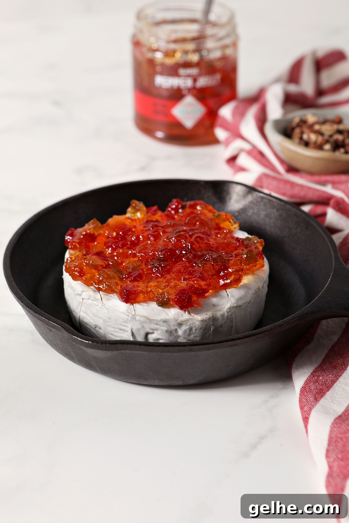 A wheel of brie in a small cast iron skillet topped with pepper jelly on a marble surface next to a red and white striped linen