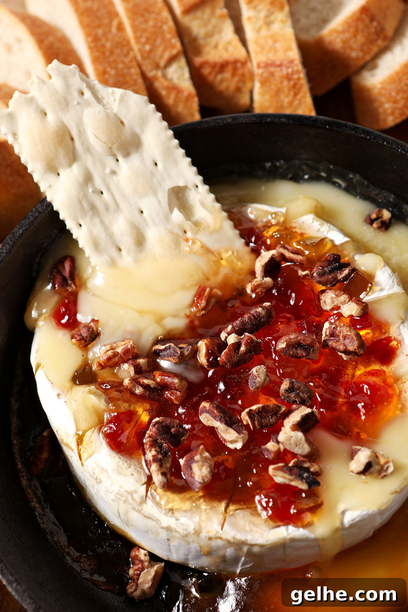 A cracker dips into gooey pepper jelly baked brie in a skillet next to baguette slices