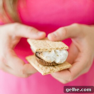 Reese’s S’mores Recipe: Homemade Chocolate Peanut Butter S’mores 9 Hands holding a smore made with two graham crackers, a roasted marshmallow, and a peanut butter cup. The person is wearing a pink shirt.