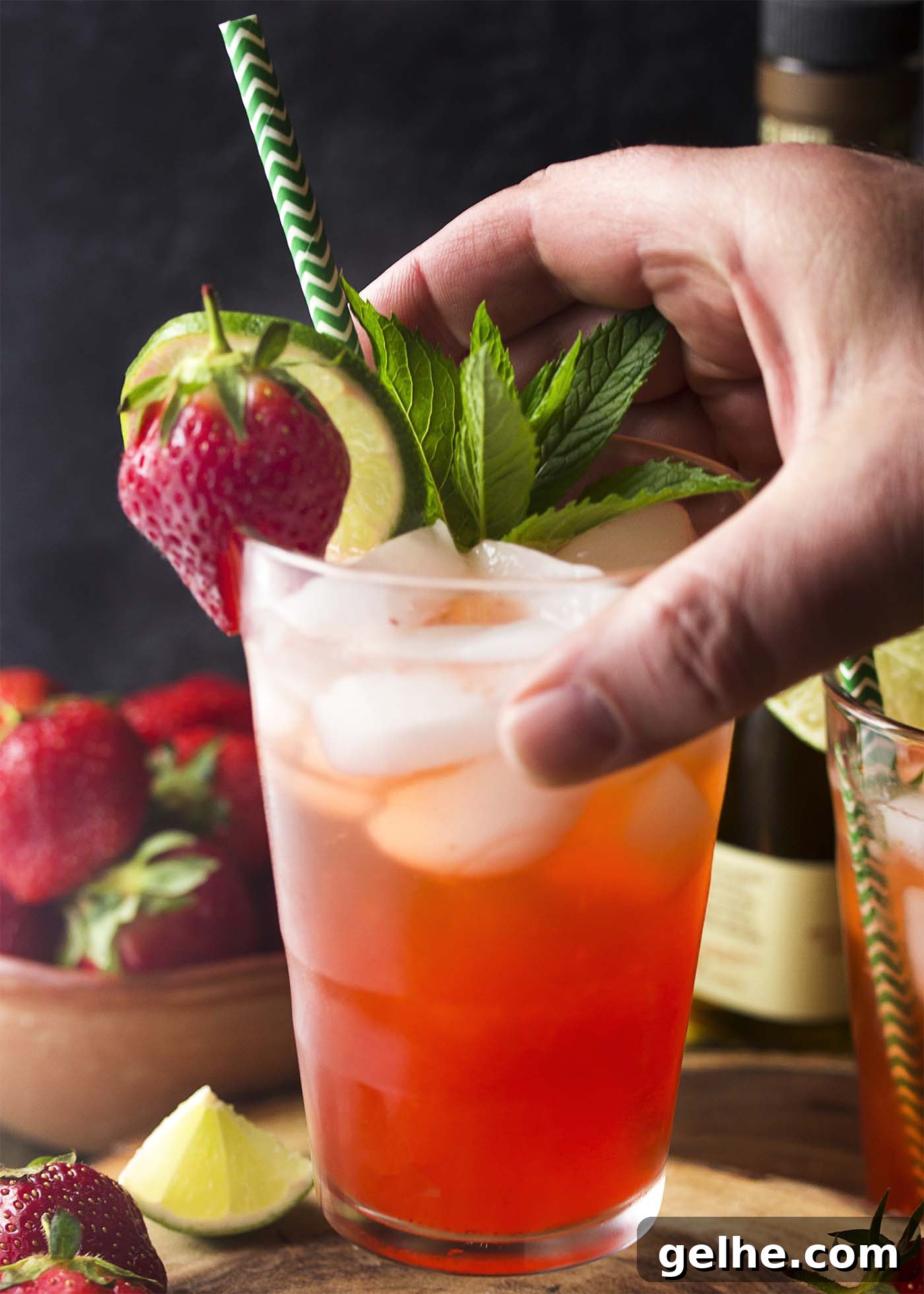 A hand lifting up a pink drink in a tall glass garnished by strawberry, lime, and mint.