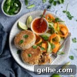 Two vegan Thai red curry fish cakes on a plate with vegetable ribbons and a bowl of sweet chilli sauce, surrounded by lime slices, sliced spring onions and fresh coriander.