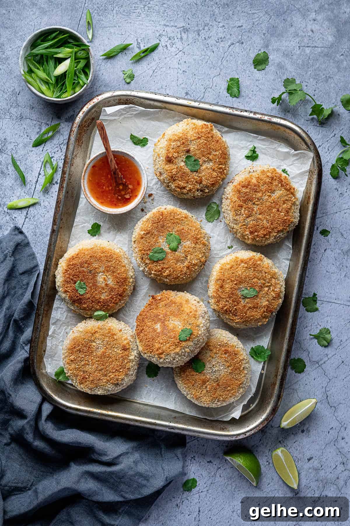 Vegan Thai red curry 'fish' cakes on a baking sheet with a bowl of sweet chilli sauce, lime slices and sliced spring onions.