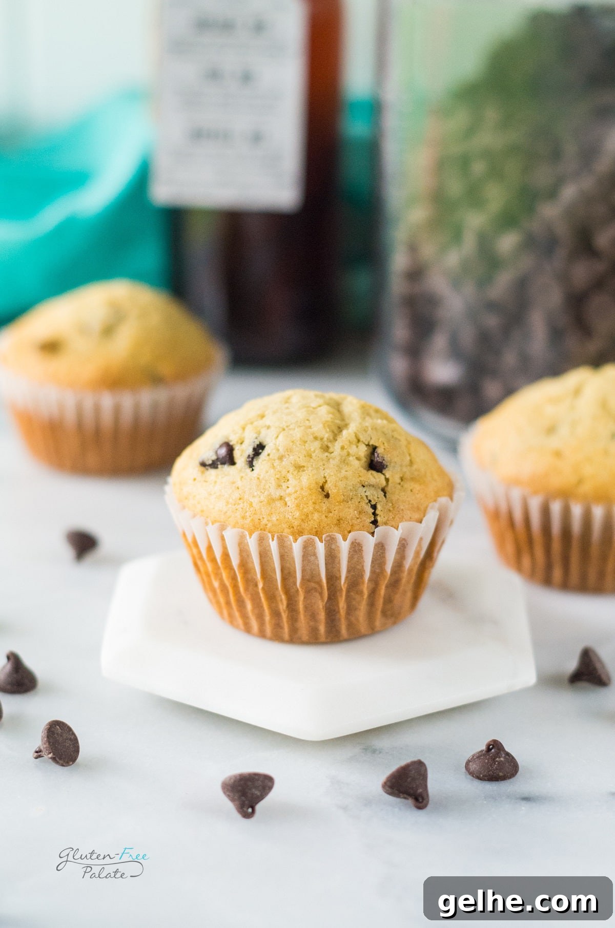 a perfectly domed gluten-free chocolate chip muffin on a white hexagon pedastal. In the background are two more muffins. Chocolate chips are scattered on the counter