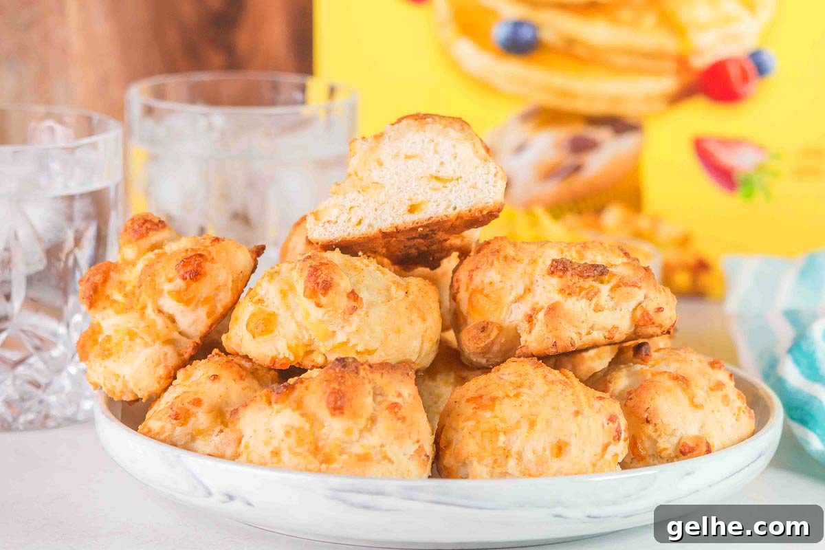 Flaky Cheddar Biscuits Made with Bisquick 2 A plate of delicious Bisquick cheddar biscuits is displayed, with one biscuit halved on top. Two glasses of water and a box with pictures of pancakes are in the background, creating a cozy and inviting scene.