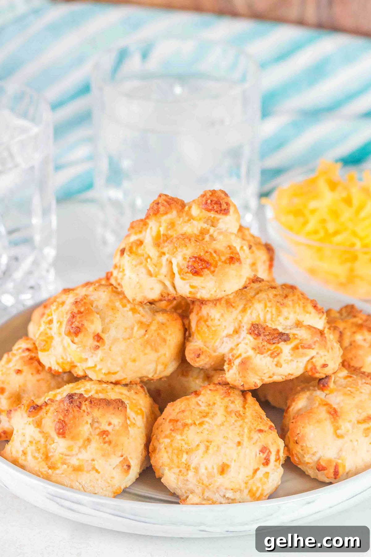Flaky Cheddar Biscuits Made with Bisquick 11 A plate of cheesy Bisquick cheddar biscuits is stacked in front of glasses with ice and a bowl of shredded cheese.