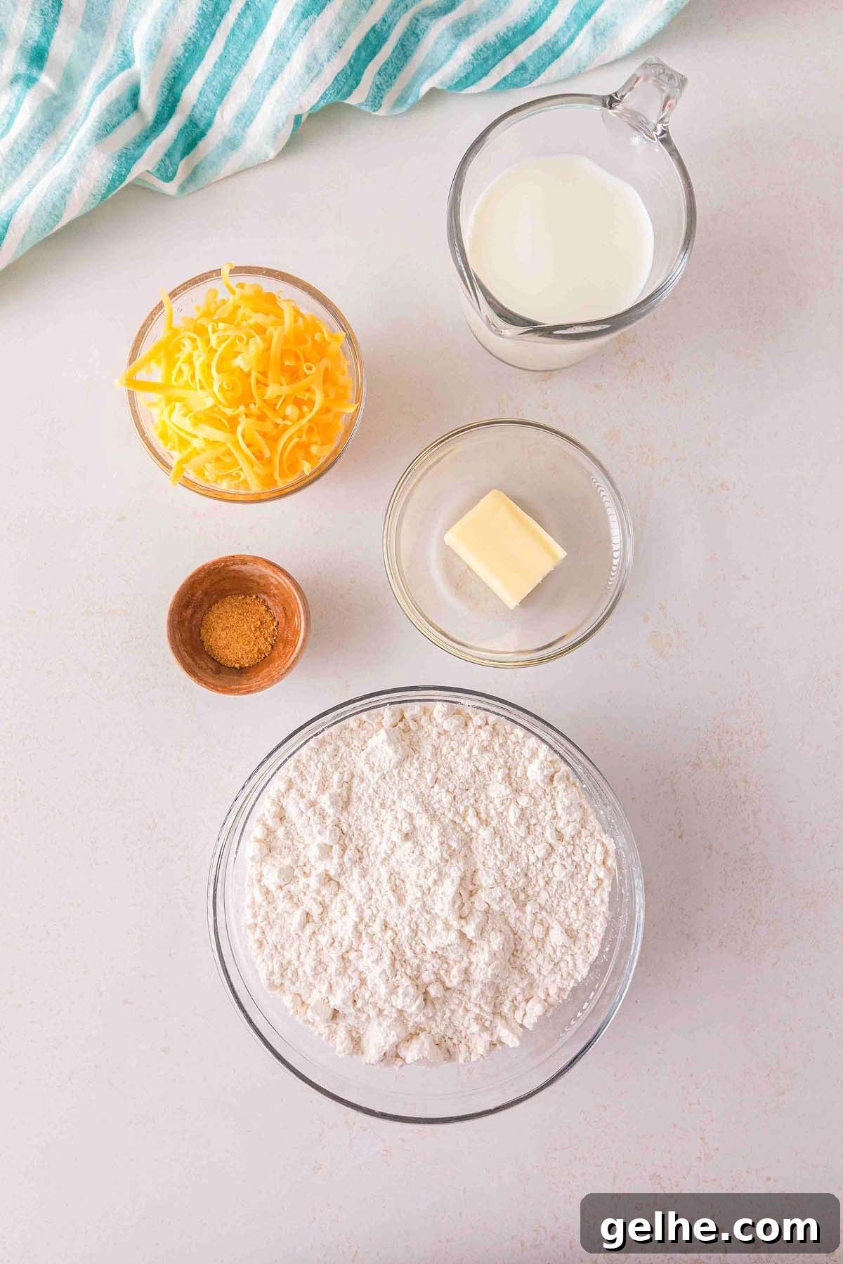 Flaky Cheddar Biscuits Made with Bisquick 13 A scene of Bisquick cheddar biscuit prep: a bowl of flour, shredded cheese, a small piece of butter, brown powder, and a pitcher of milk sit ready on the surface. A striped cloth rests in the corner.