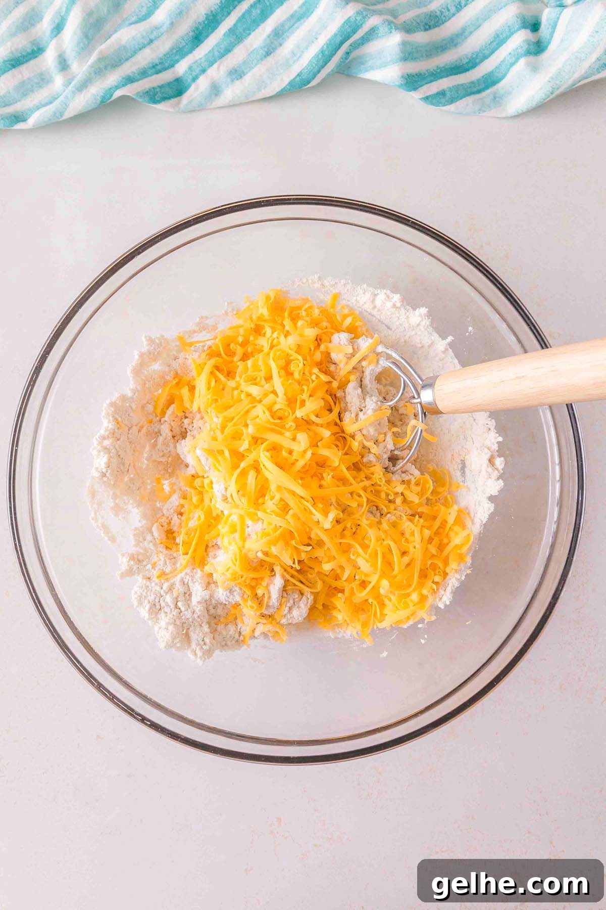 Flaky Cheddar Biscuits Made with Bisquick 14 A glass bowl with a Bisquick cheddar biscuit mix of shredded cheese and flour, accompanied by a wooden-handled whisk, sits on a light countertop. A striped cloth is partially visible in the top corner.