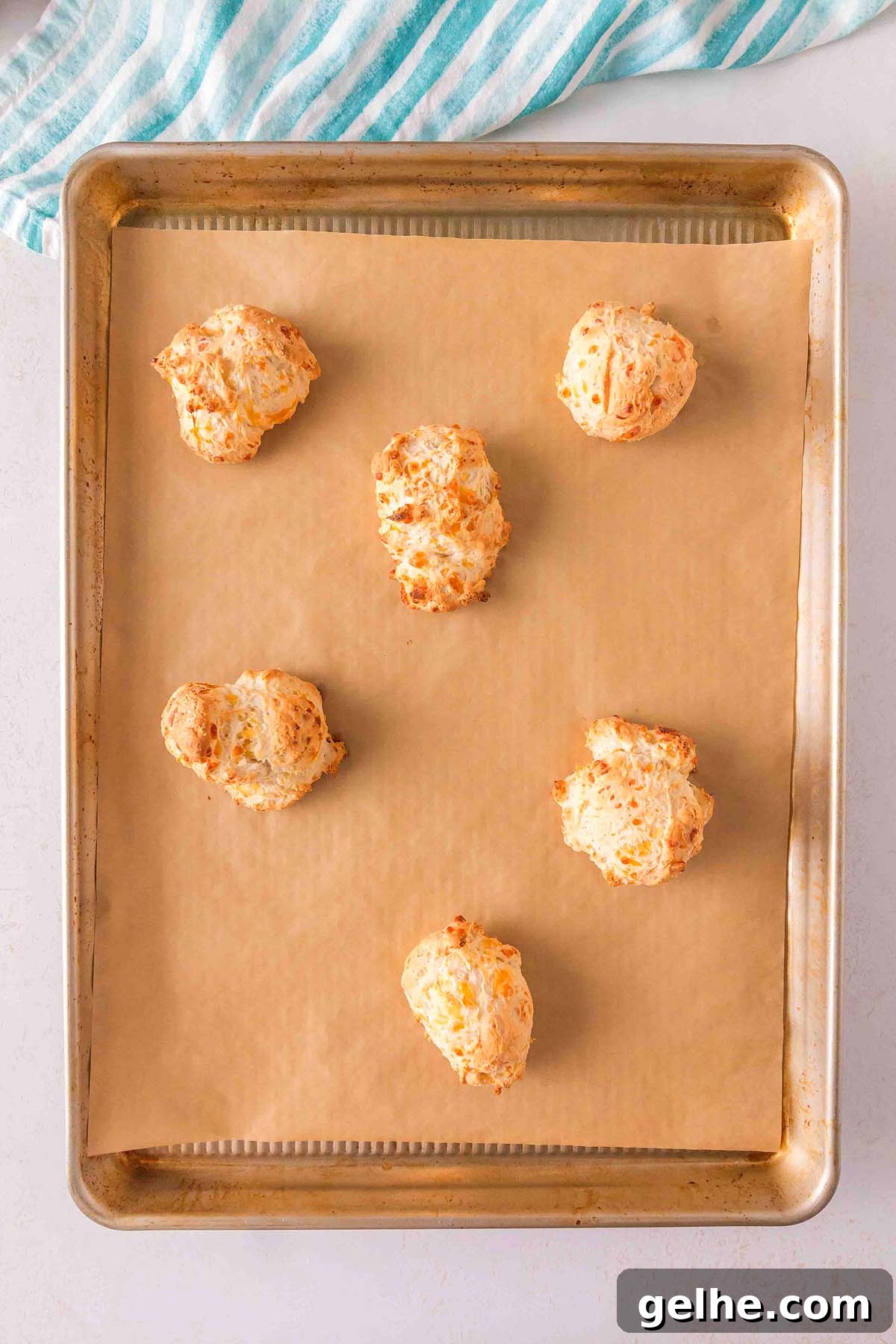 Flaky Cheddar Biscuits Made with Bisquick 17 Six baked cheese puffs, reminiscent of Bisquick cheddar biscuits, rest on a parchment-lined baking sheet. A striped cloth is partially visible at the top.