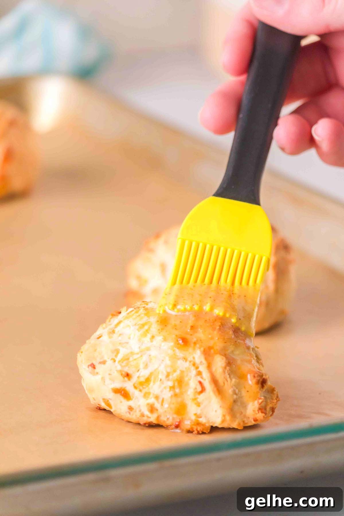 Flaky Cheddar Biscuits Made with Bisquick 18 A hand uses a yellow brush to glaze freshly baked Bisquick cheddar biscuits on a parchment-lined baking sheet.