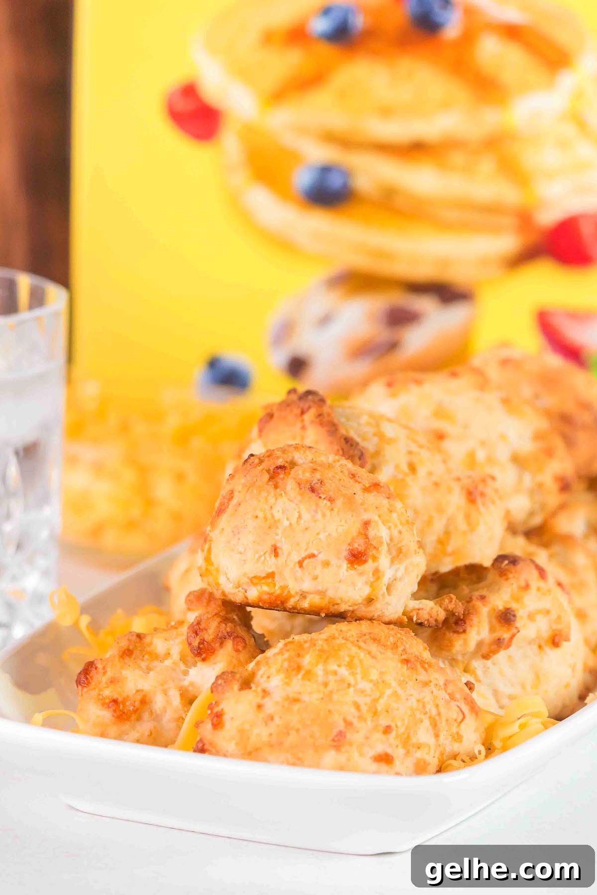 Flaky Cheddar Biscuits Made with Bisquick 9 A plate of Bisquick cheddar biscuits rests on a white dish, accompanied by a refreshing glass of water. In the background, a box adorned with pancake imagery hints at more delightful possibilities.