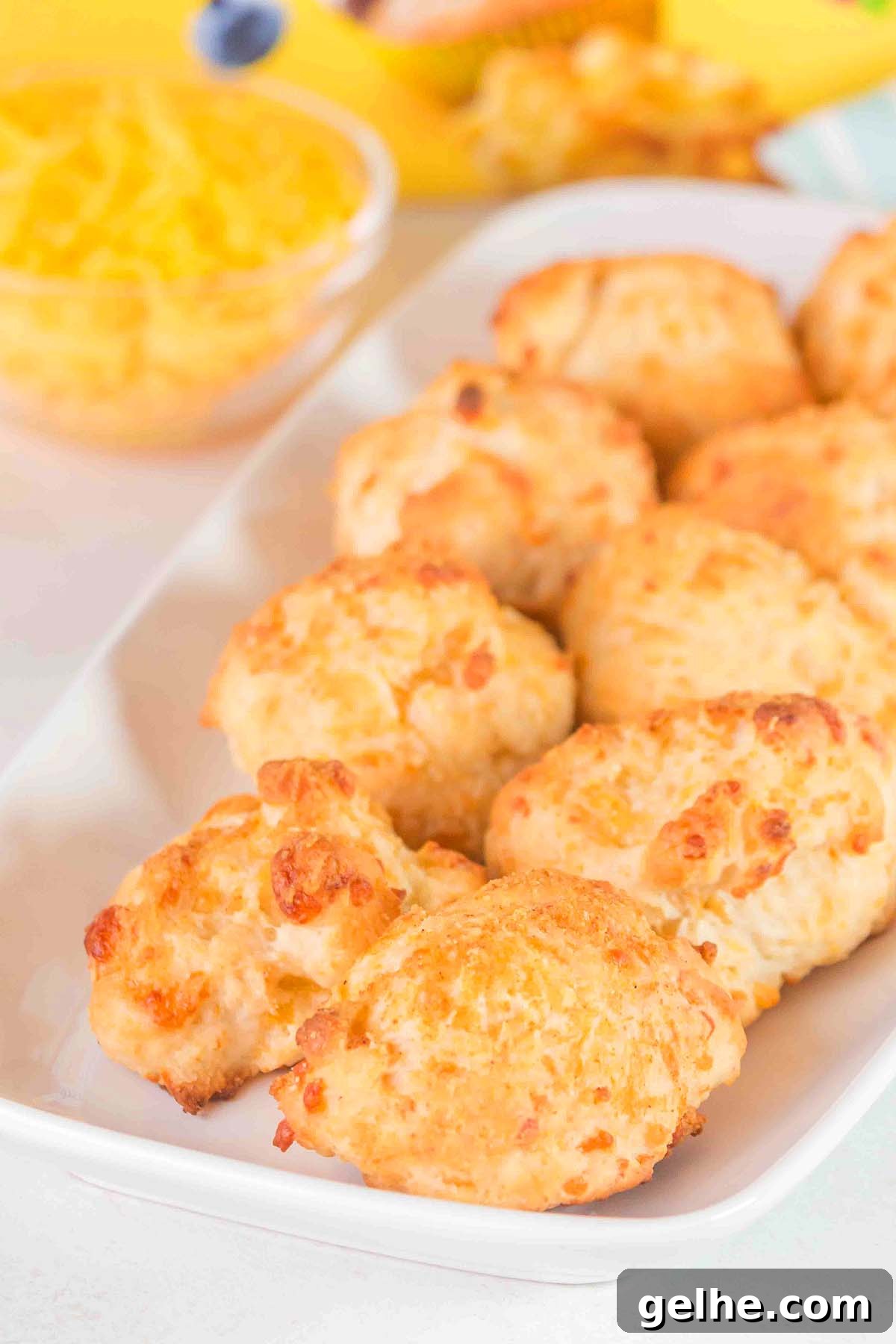Flaky Cheddar Biscuits Made with Bisquick 10 A white plate with a stack of golden-brown Bisquick cheddar biscuits rests next to a bowl of grated cheese on a white surface.