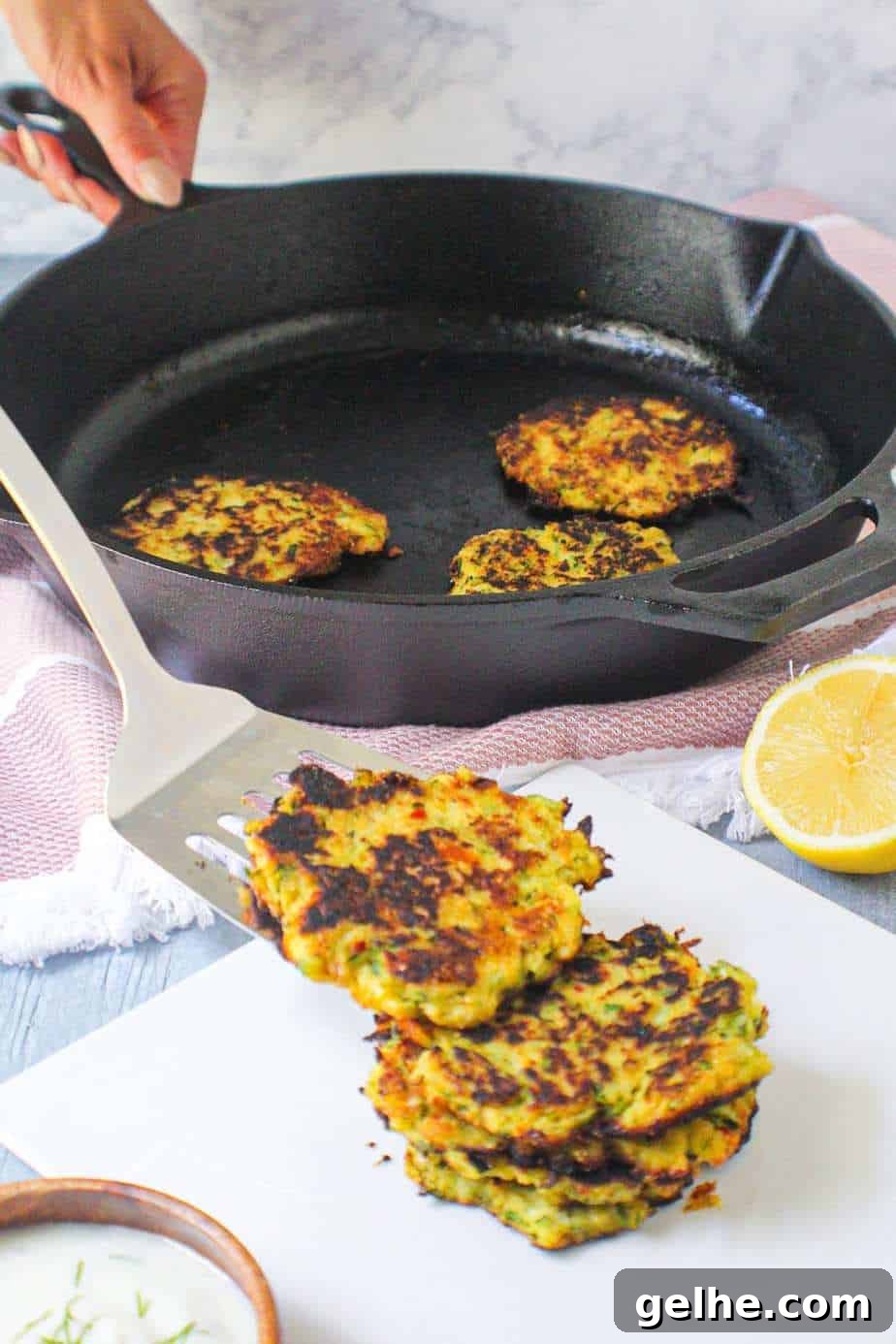 Crispy Cornmeal Zucchini Fritters with Garlic Herb Dip 3 side angle shot of cornmeal zucchini fritters being placed on a white plate with a cast iron skillet of fritters in the background.