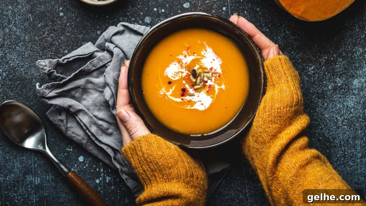 Female hands in yellow knitted sweater holding a bowl with pumpkin cream soup on dark stone background with spoon decorated with cut fresh pumpkin, top view. Autumn cozy dinner concept 
