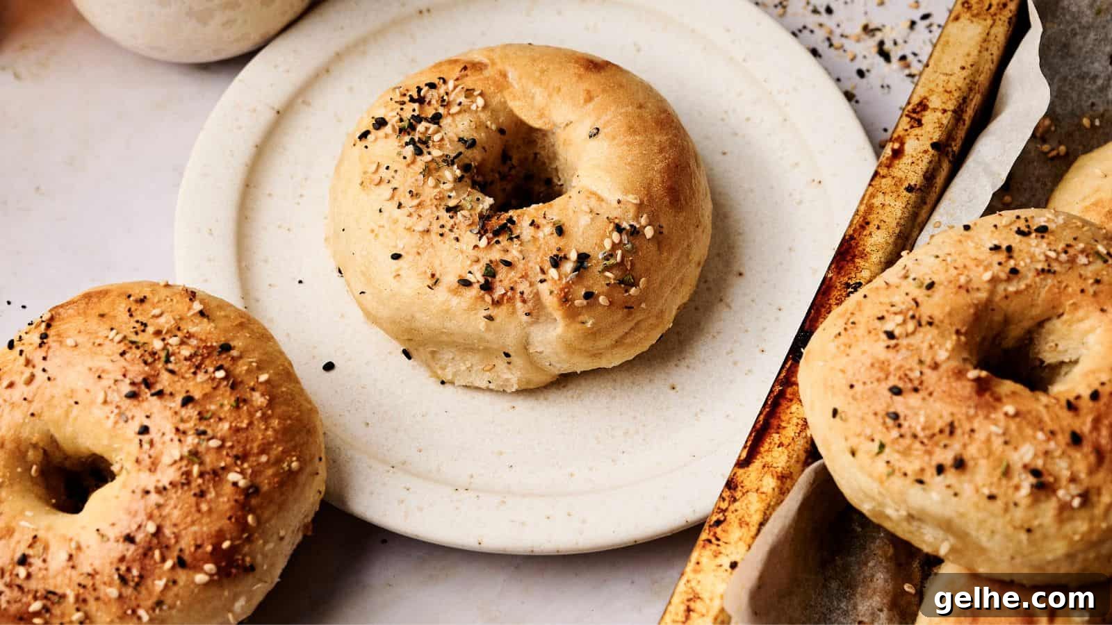 A plate with a bagel seasoned with seeds sits next to a baking tray holding more bagels, also topped with seeds.