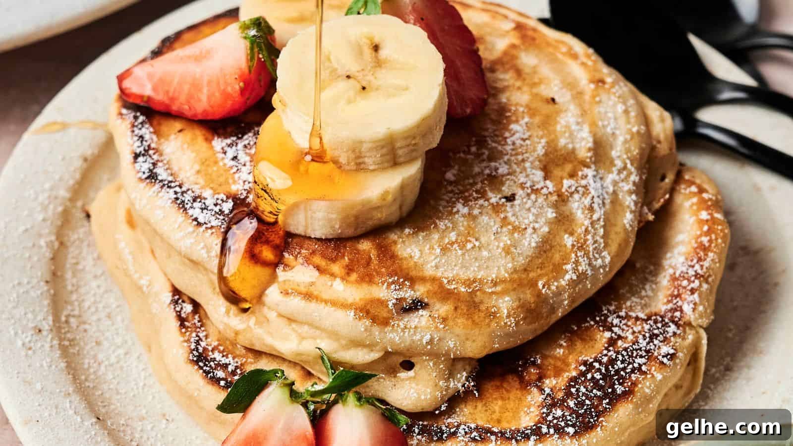 A stack of pancakes topped with sliced bananas, strawberries, and syrup on a plate, accompanied by black utensils.