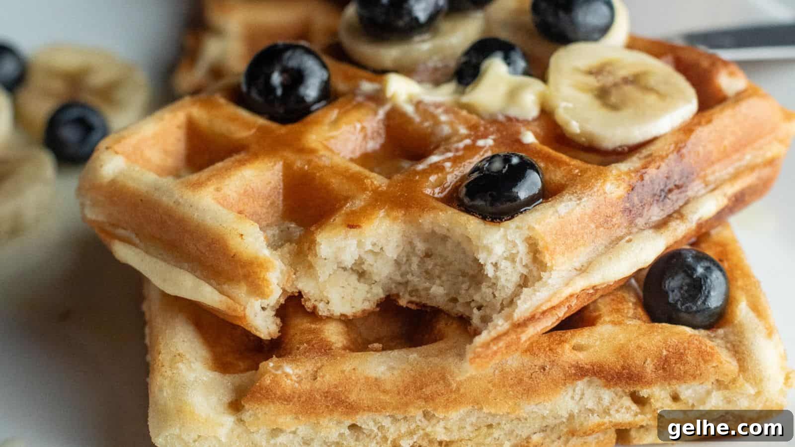 Two stacked waffles with a bite taken out, topped with banana slices, blueberries, and a pat of butter.