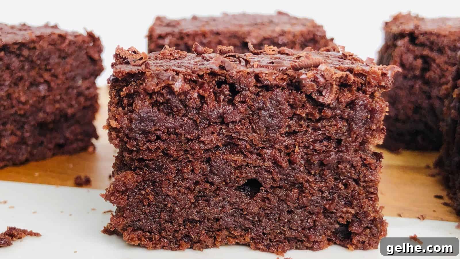 Close-up of a square cut chocolate brownie with a rich, dense texture, resting on a light-colored surface. More brownies are visible in the background.