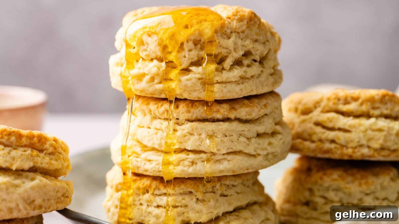 A stack of flaky biscuits with honey drizzled on top, set on a plate with more biscuits in the background.