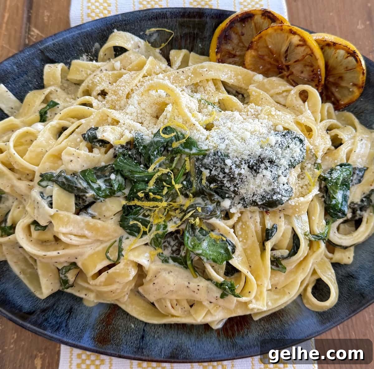 Creamy lemon pasta with spinach in blue serving platter with sliced lemons and lemon zest garnish and sprinkled with grated pecorino cheese.