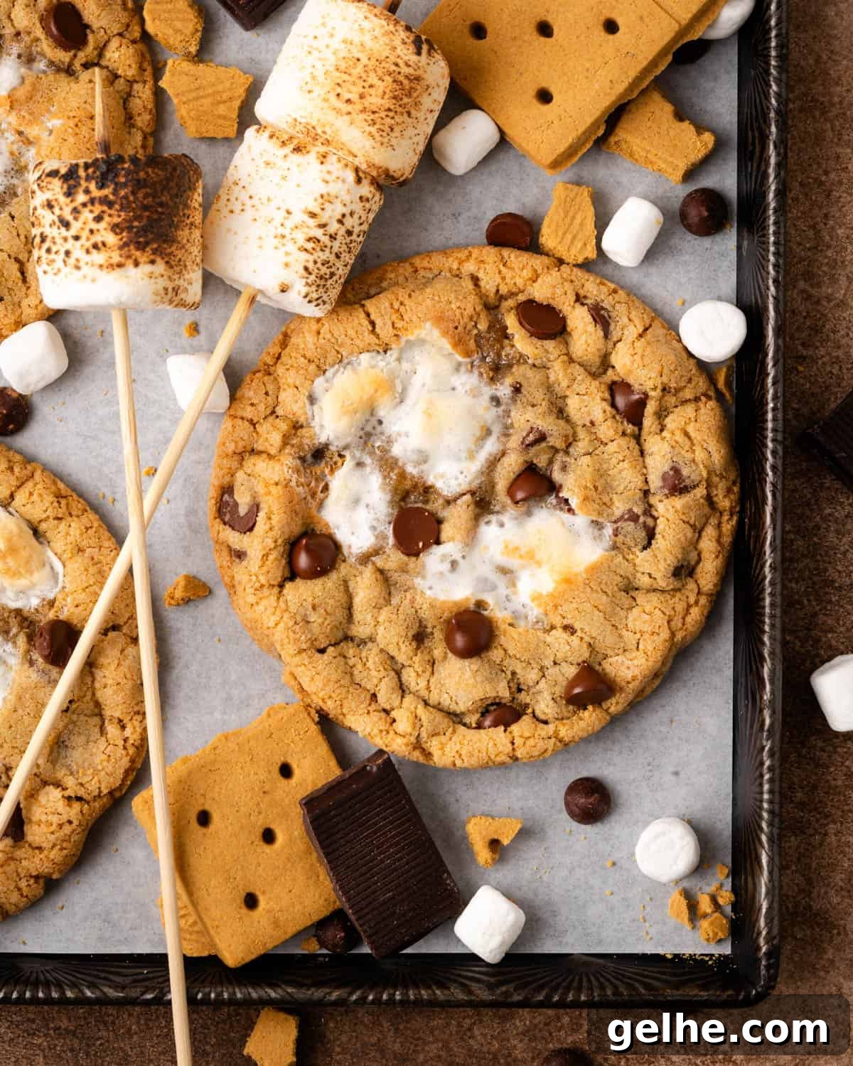 S'mores chocolate chip cookies laying on a baking sheet surrounded by graham cracker pieces, chocolate bars and toasted marshmallows.