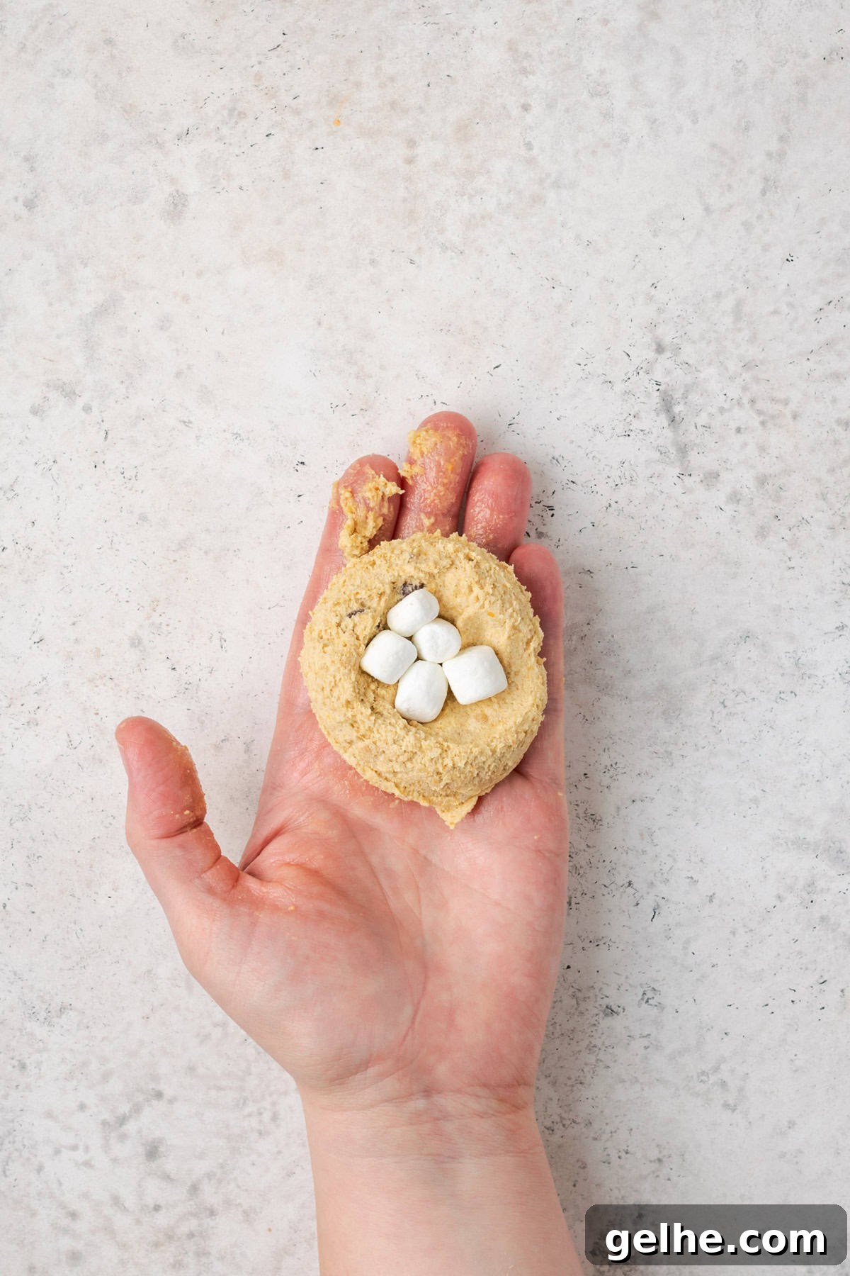 A hand holding a large piece of cookie dough that has five mini marshmallows pressed into the center.