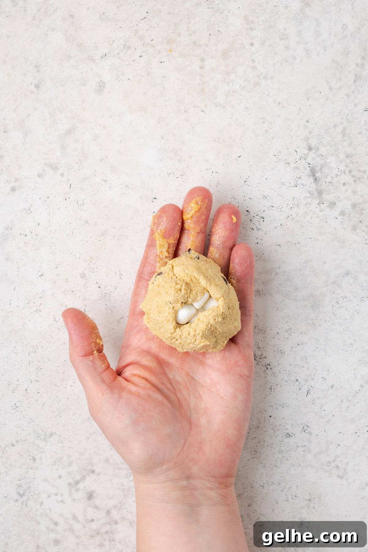 A hand holding a cookie dough ball, where the dough has been wrapped around the marshmallow center.
