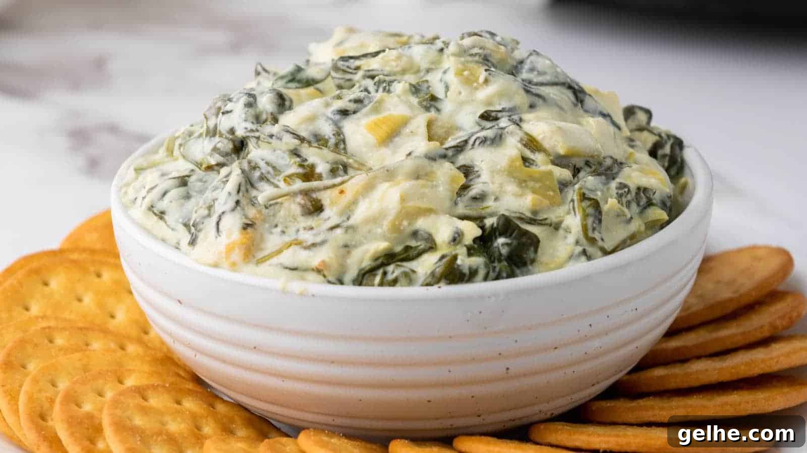 21 Labor Day Picnic Recipes for a Crowd: Ultimate Menu Ideas 9 A white bowl of creamy spinach artichoke dip surrounded by round crackers on a marble surface.
