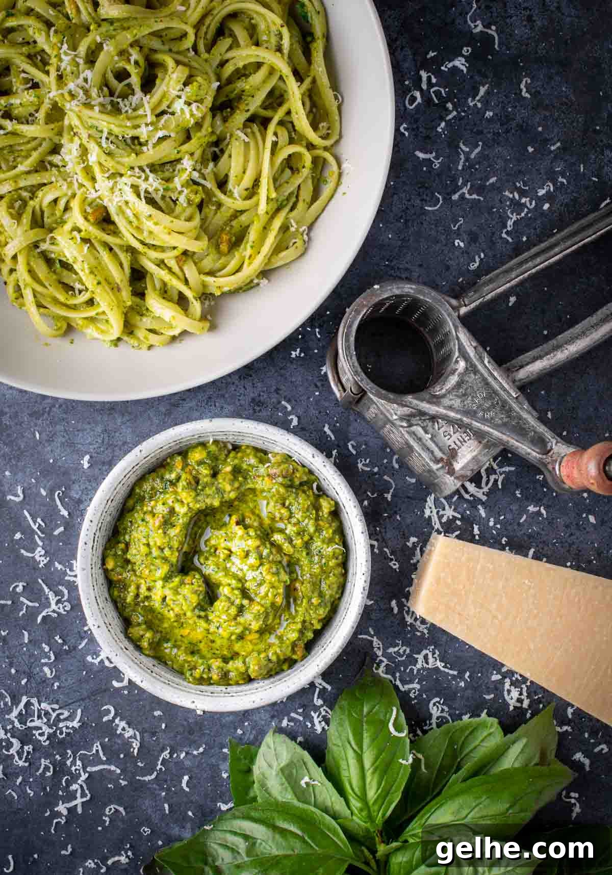 A bowl of pistachio peso surrounded by a bowl of pasta, a parmesan grater and parmesan cheese.