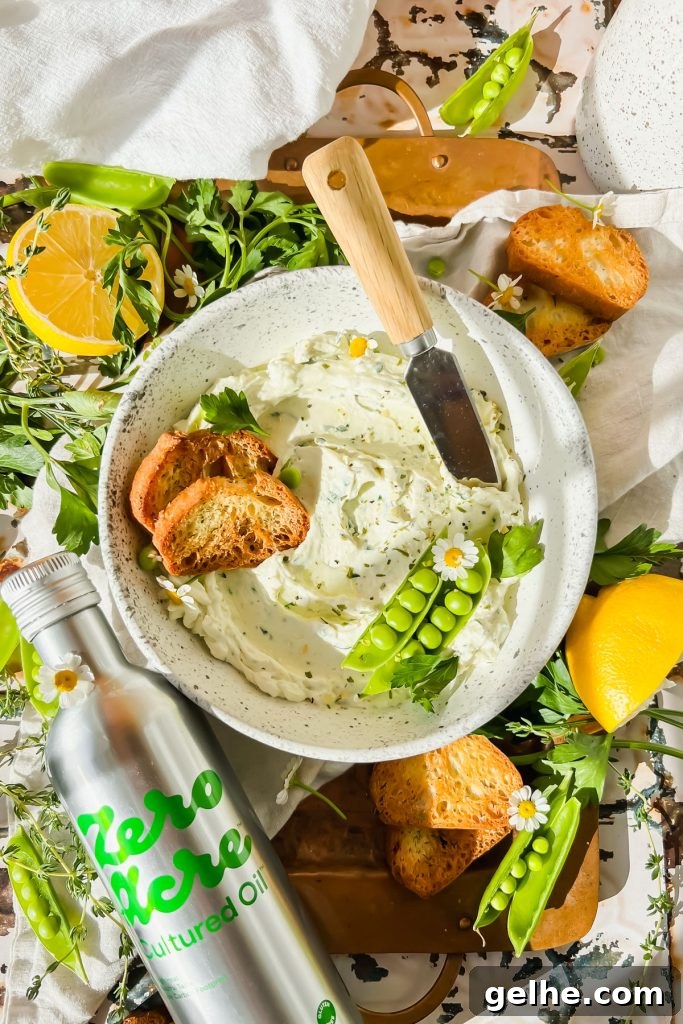 Baked Cheddar Jalapeño Dip with Crispy Panko Topping 11 cream cheese garlic dip in white bowl with two toasted crostinis and pea pods in bowl beside bottle of zero acre cultured oil with fresh herbs surrounding.