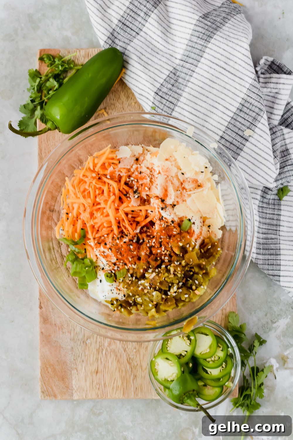 Baked Cheddar Jalapeño Dip with Crispy Panko Topping 3 ingredients for cheddar jalapeño dip unmixed in glass bowl on top of wood cutting board beside small bowel of jalapeño slices and striped tea towel.