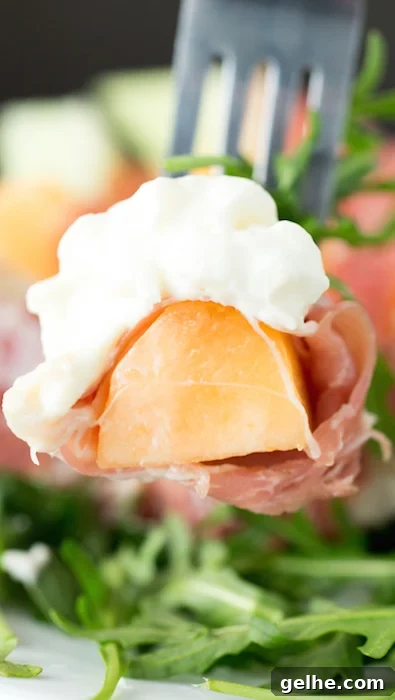 Prosciutto, Melon & Burrata Salad with Honey-Lemon Dressing 4 A fork holding a bite sized piece of cantaloupe that's wrapped in prosciutto and has a bit of burrata on top.