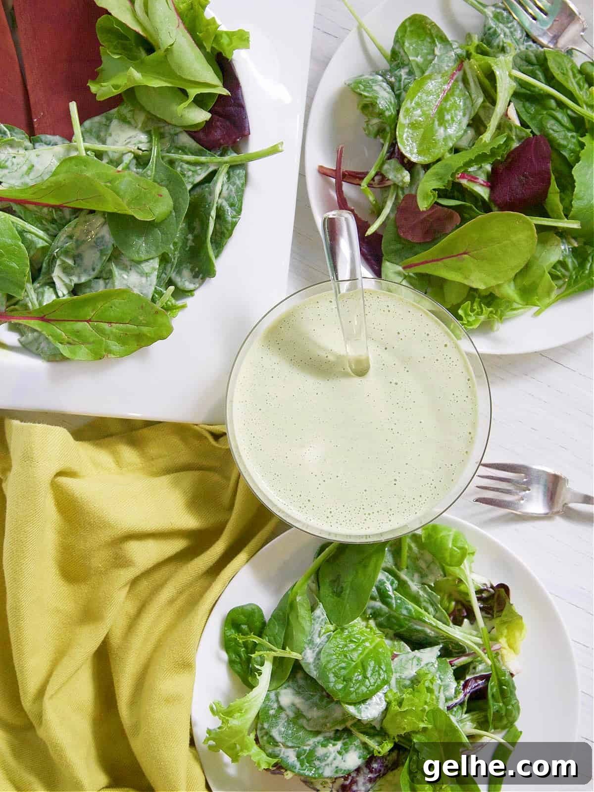Two white plates with salad green tossed in green goddess dressing and silver forks.