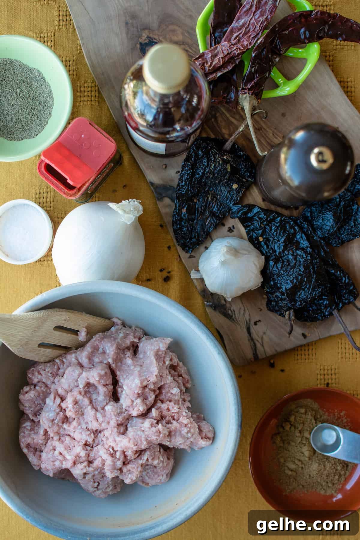 Ingredients to make chorizo on a table with a yellow tablecloth. Ground pork in a bowl alongside an onion, dried chiles, and spices.