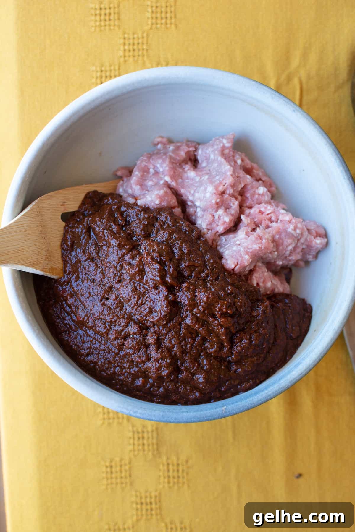 A ceramic bowl filled with ground pork and red Chile paste with a wooden spoon sticking out of one side of the bowl.