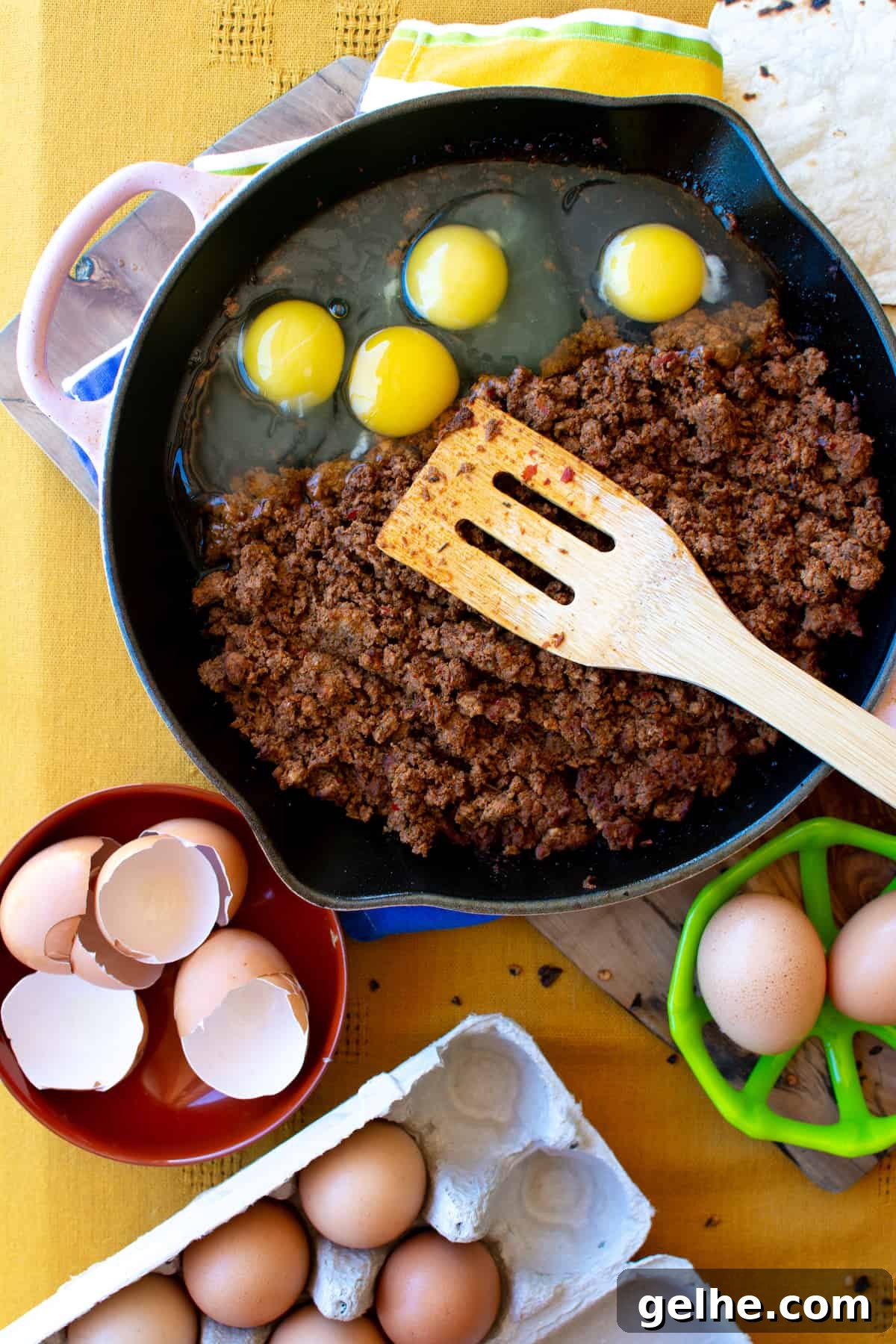 A skillet with cooked chorizo and four raw eggs with a wooden spoon sitting on top of the chorizo in the skillet.