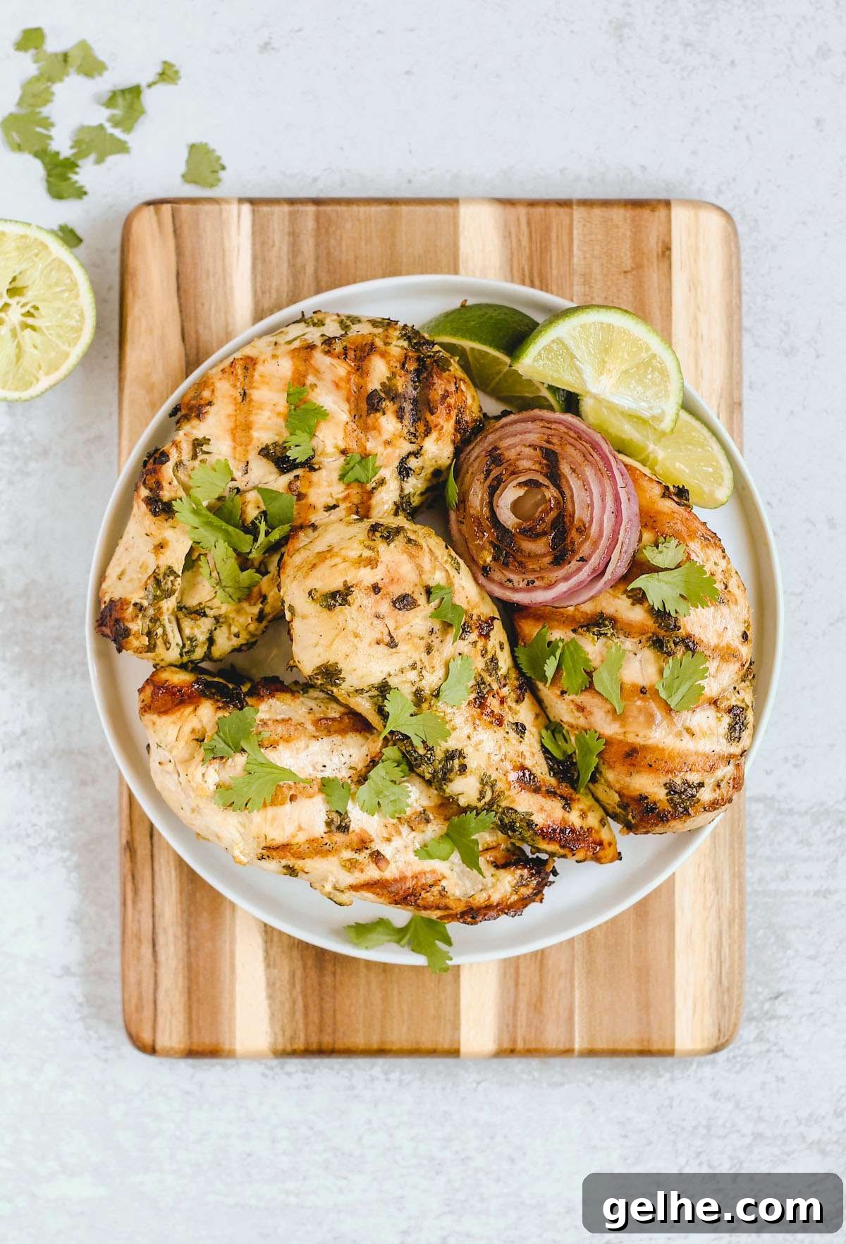 Four grilled cilantro lime chicken breasts with grilled onions and lime slices on a round plate on top of a wood cutting board.