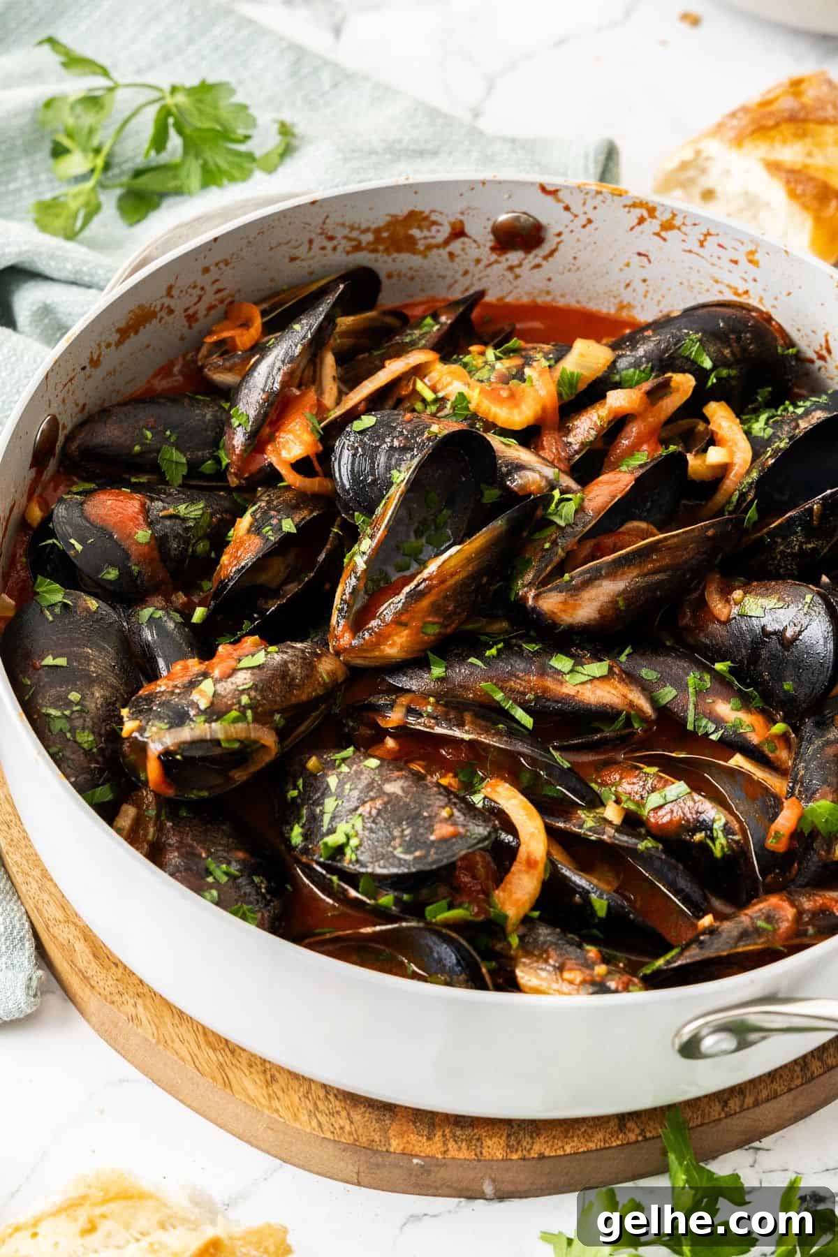 Frying pan with steamed mussels in tomato sauce, sprinkled with a parsley garnish, and some pieces of bread around the edge.