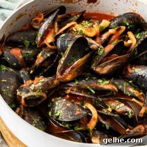 Frying pan with steamed mussels in tomato sauce, sprinkled with a parsley garnish, and some pieces of bread around the edge.