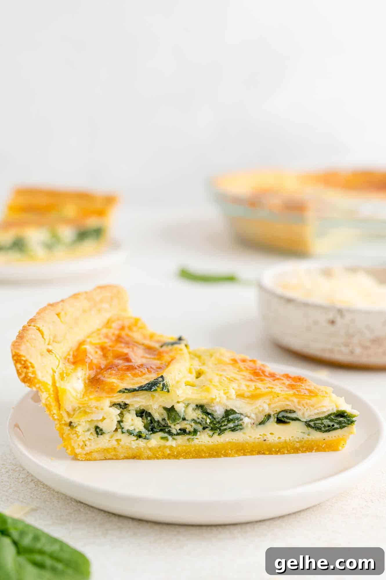 Slice of quiche on a white plate, more quiche in background.