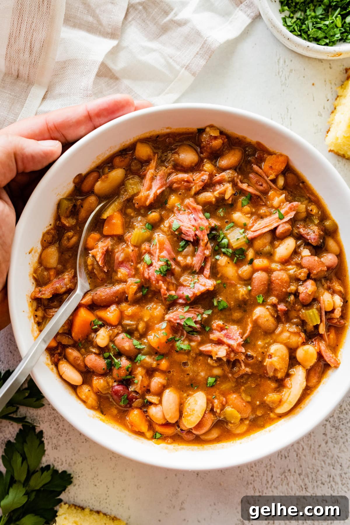 15 bean soup mix recipe in a white bowl with slices of cornbread on the side. Parsley for garnish. close up of bowl and soup.