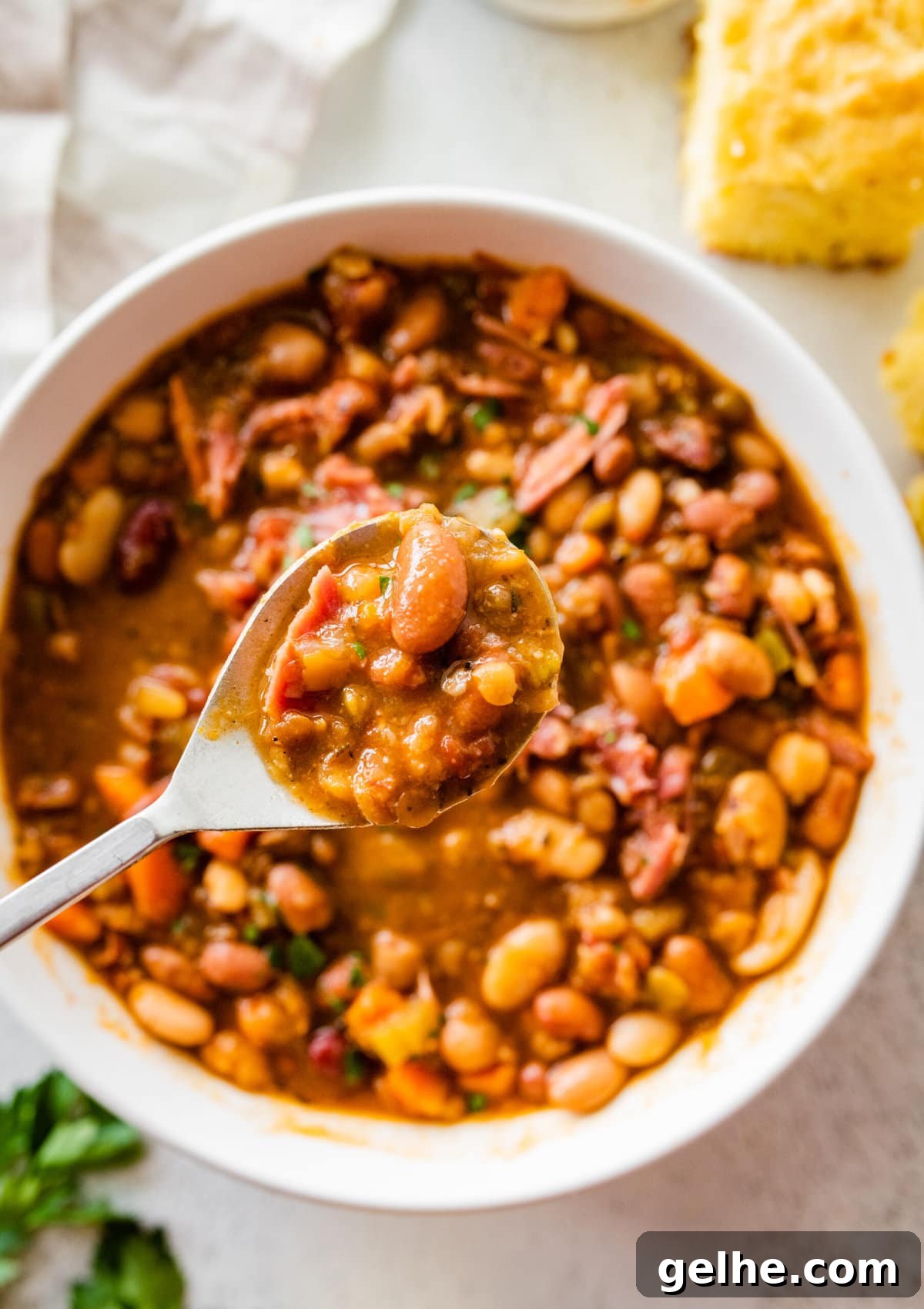 15 bean soup mix recipe in a white bowl with slices of cornbread on the side. Parsley for garnish. Spoon showing the soup.