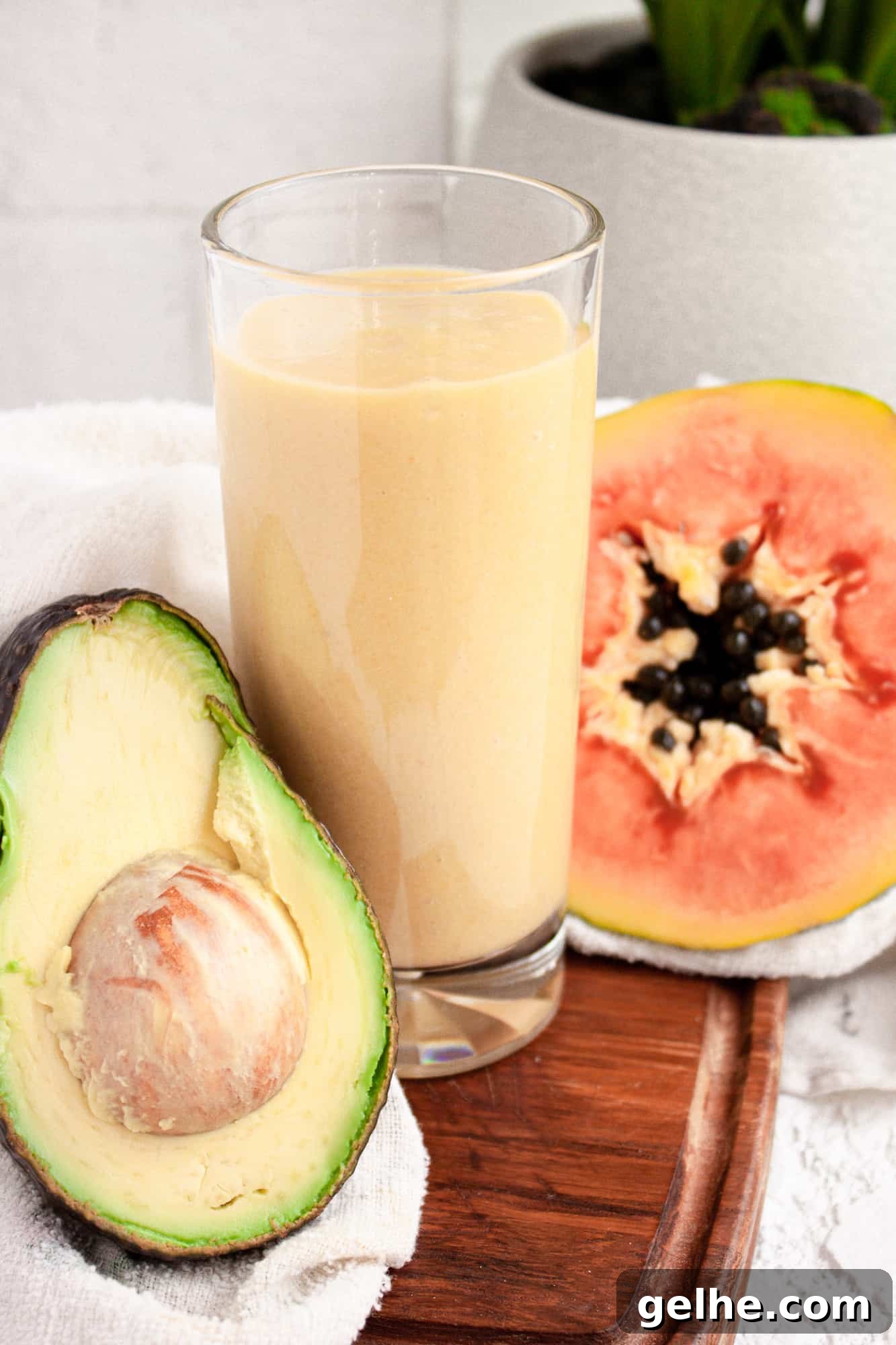 Creamy Papaya Avocado Smoothie Recipe for Glowing Skin 3 Up close view of a glass of papaya avocado smoothie on a wooden board beside half an avocado and papaya
