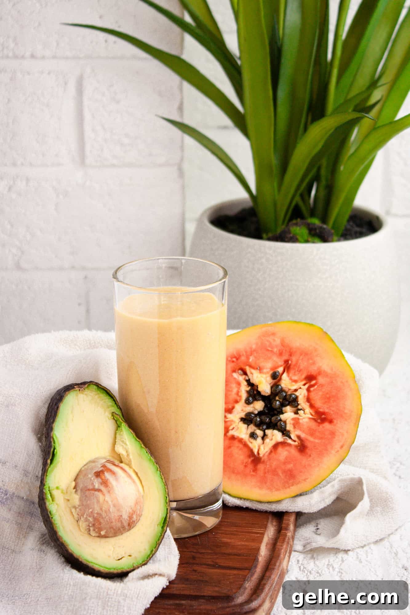Creamy Papaya Avocado Smoothie Recipe for Glowing Skin 4 Tall glass of papaya avocado smoothie on a wooden board beside half an avocado and papaya, in front of a white brick wall
