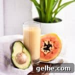 Creamy Papaya Avocado Smoothie Recipe for Glowing Skin 5 Tall glass of papaya avocado smoothie on a wooden board beside half an avocado and papaya, in front of a white brick wall