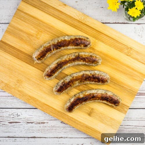 Four sous vide sausages are arranged in a diagonal line on a wooden cutting board, with a small vase of yellow flowers in the top right corner.