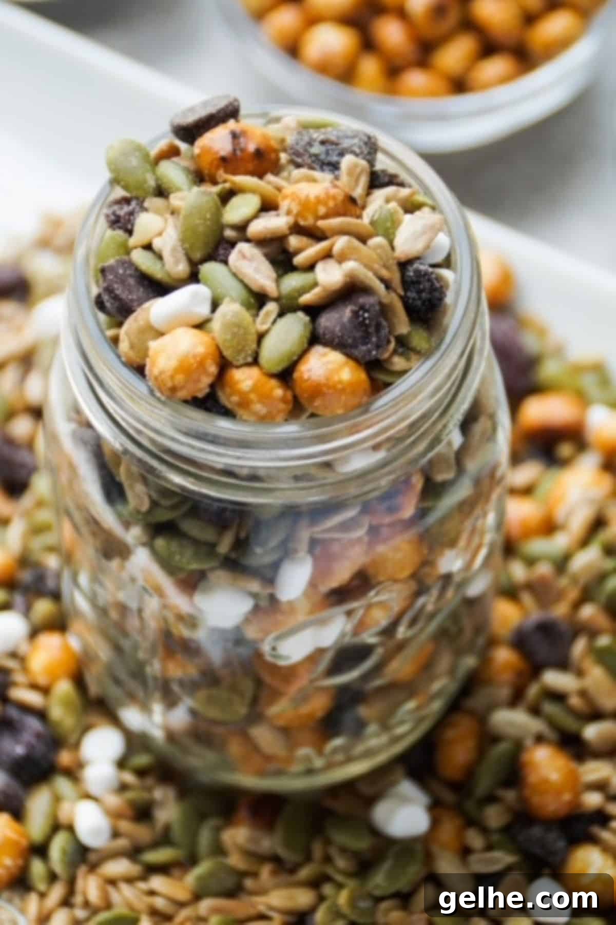 One glass mason jar filled with peanut-free trail mix. Base of jar surrounded by trail mix on a white plate. White bowl with pretzel balls partially pictured in the background.