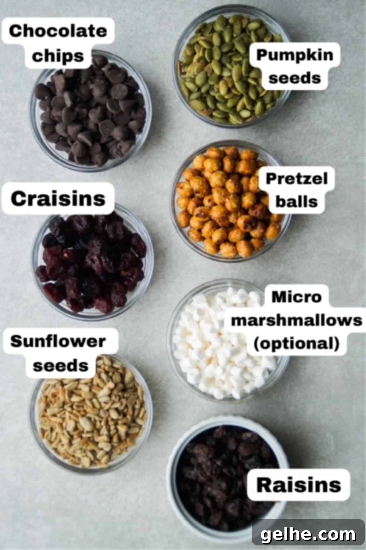 Trail mix ingredients in individual small glass bowls lined up into rows, each are labeled: Chocolate chips, craisins, sunflower seeds, pumpkin seeds, pretzel balls, micro marshmallows, and raisins.