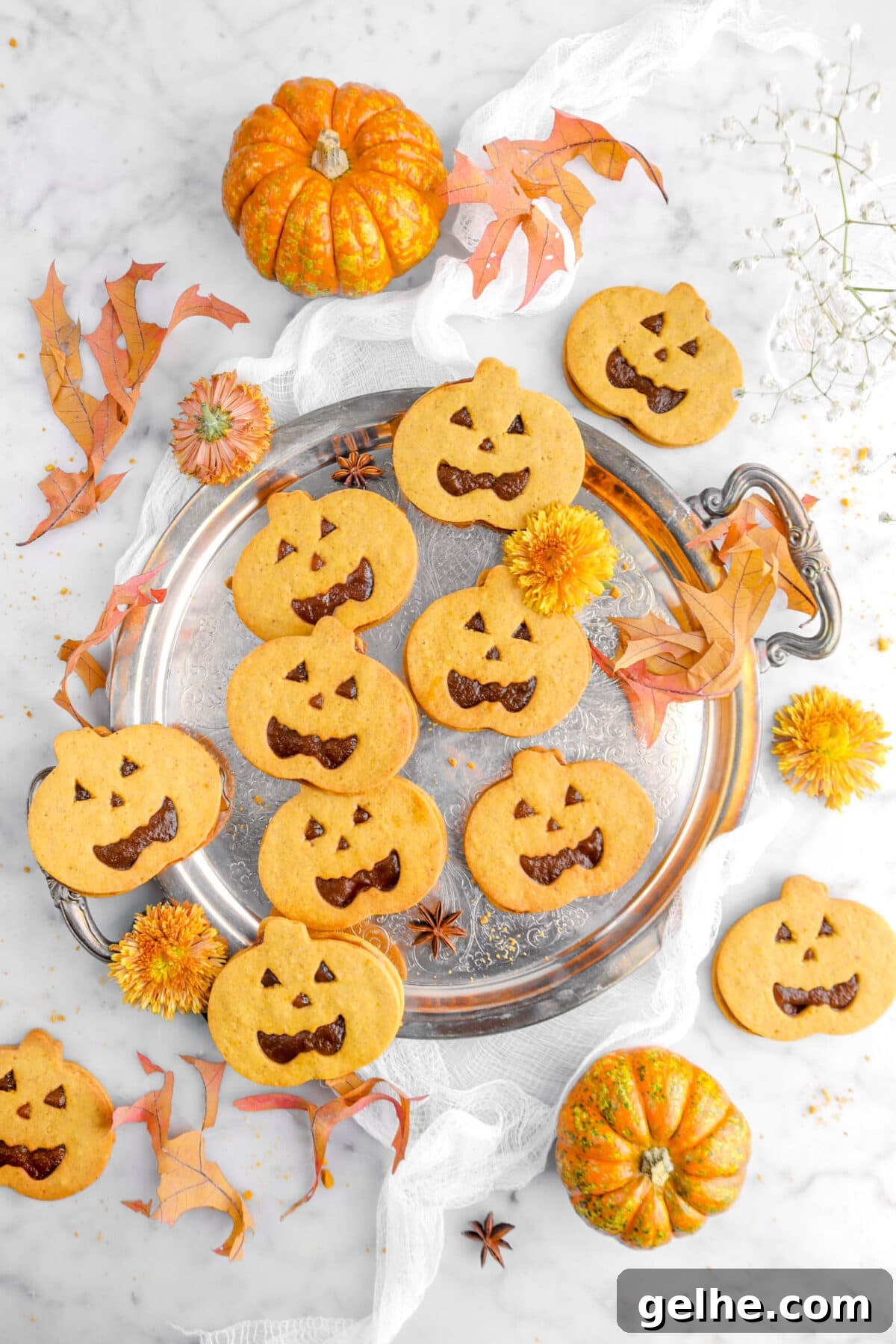 overhead shot of pumpkin jack o'lantern sandwich cookies on silver tray with leaves, yellow chrysanthemums, and orange pumpkins around on marble surface.